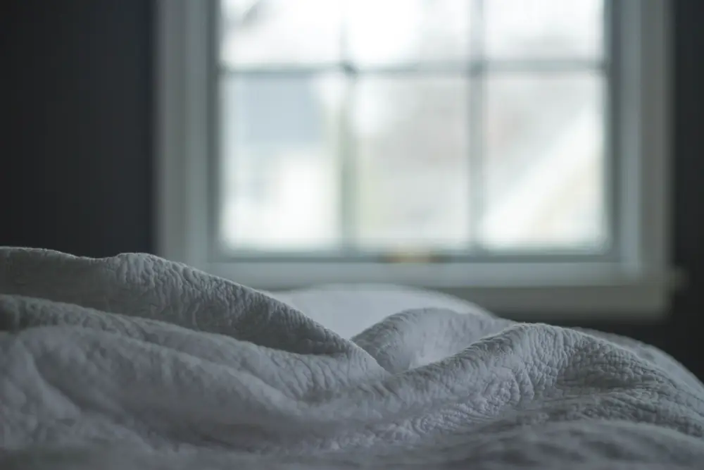 A quilted blanket on a bed beside a window in soft morning light