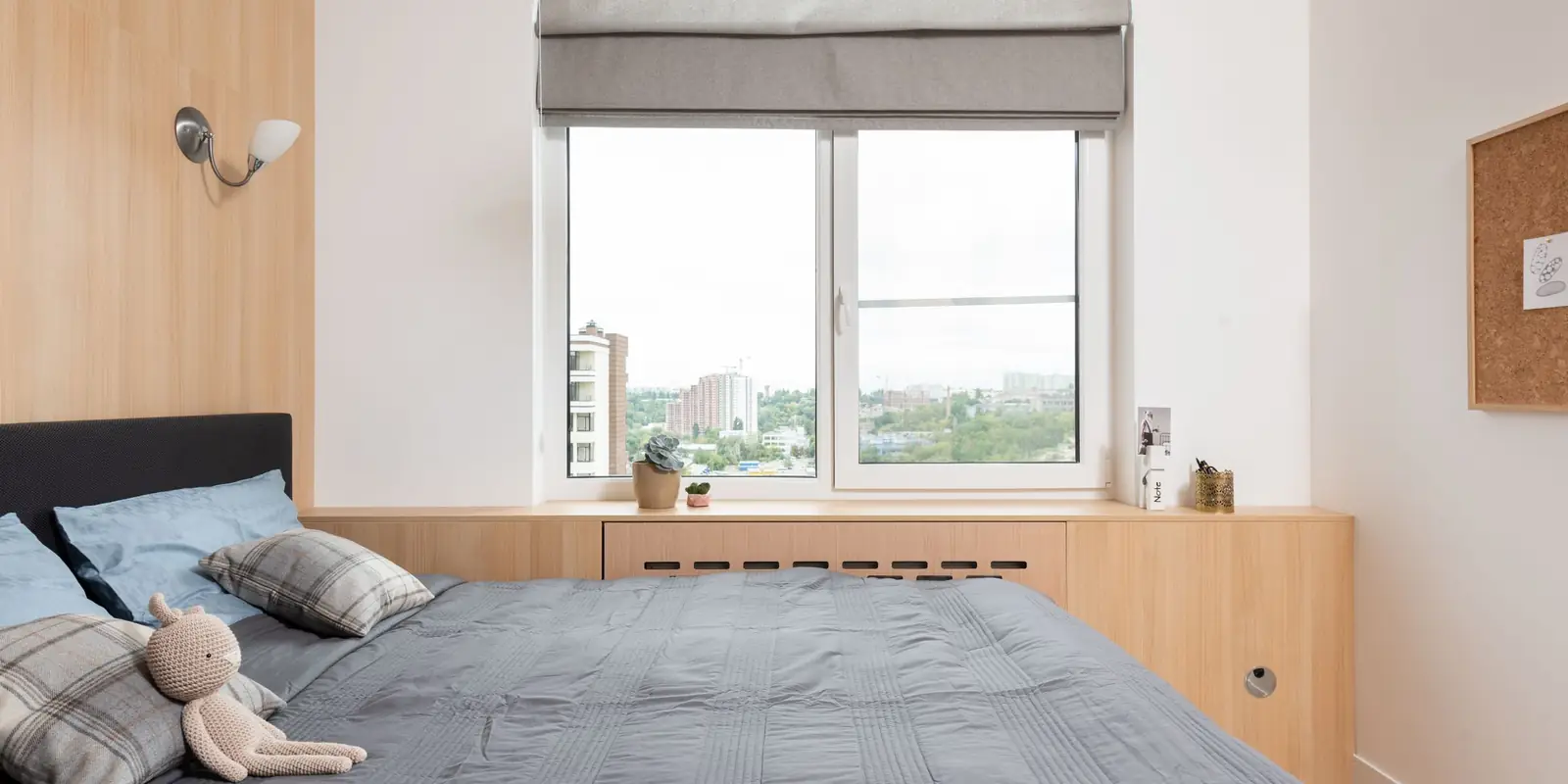 A neatly made bed with pillows and a soft toy beside a window in a small, bright room