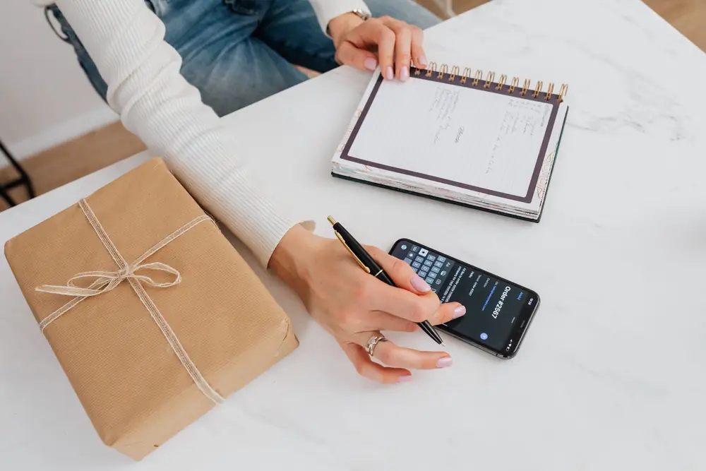 Person writing a list in a notebook beside a phone and a wrapped parcel