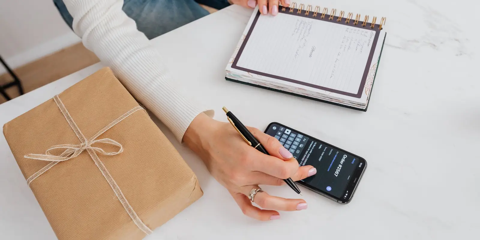 Person writing a shopping list in a notebook beside a phone and a parcel wrapped in brown paper