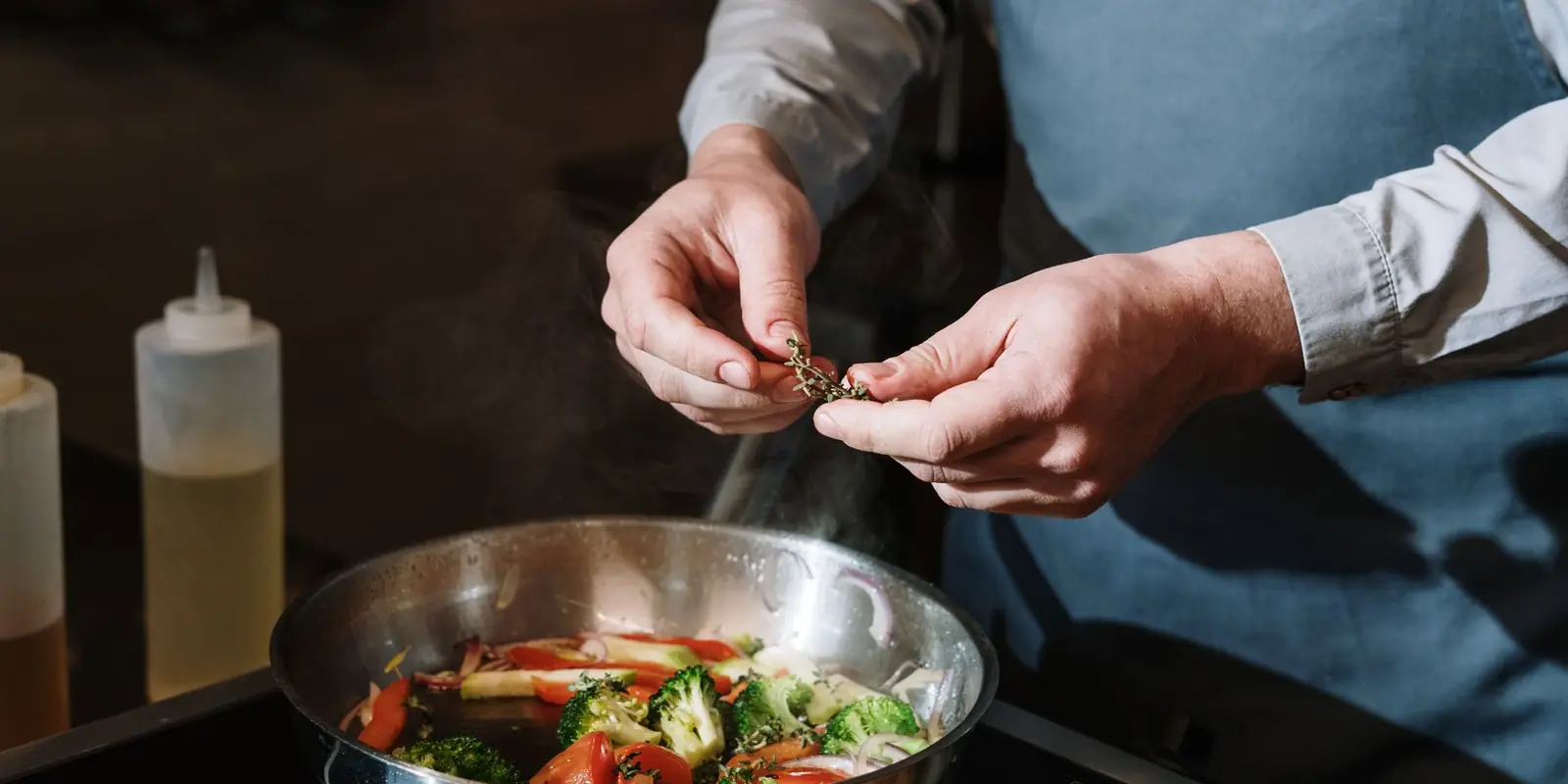 Cook in an apron adding herbs to a pan of vegetables on the hob