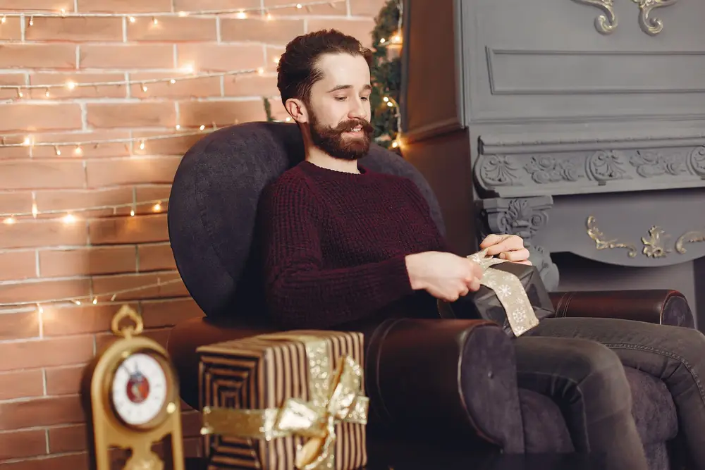 Man in an armchair opening a Christmas present beside a fireplace