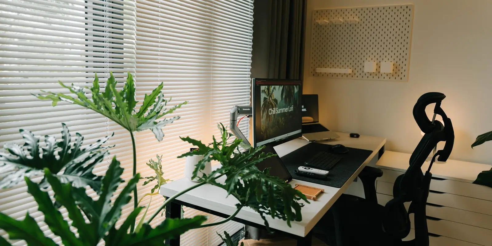 Home office desk with a monitor and office chair beside leafy plants and a window blind
