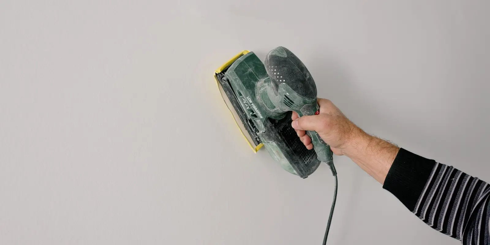 A hand sanding a white wall with a small power sander