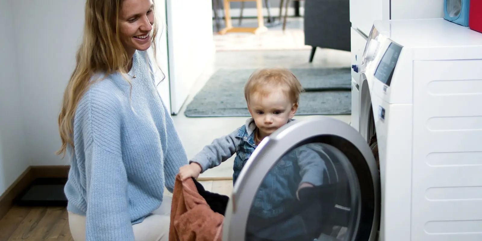 Smiling woman and a toddler unloading laundry from a front-loading machine at home