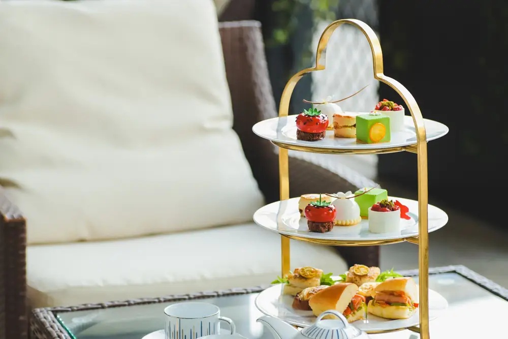A three-tier afternoon tea stand with sandwiches and small cakes beside a teapot