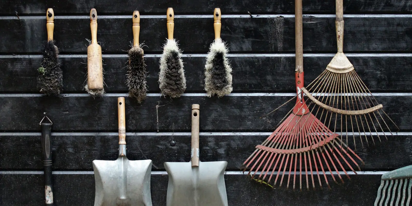 Old garden brushes, trowels and rakes hanging in a row on a dark wooden shed wall