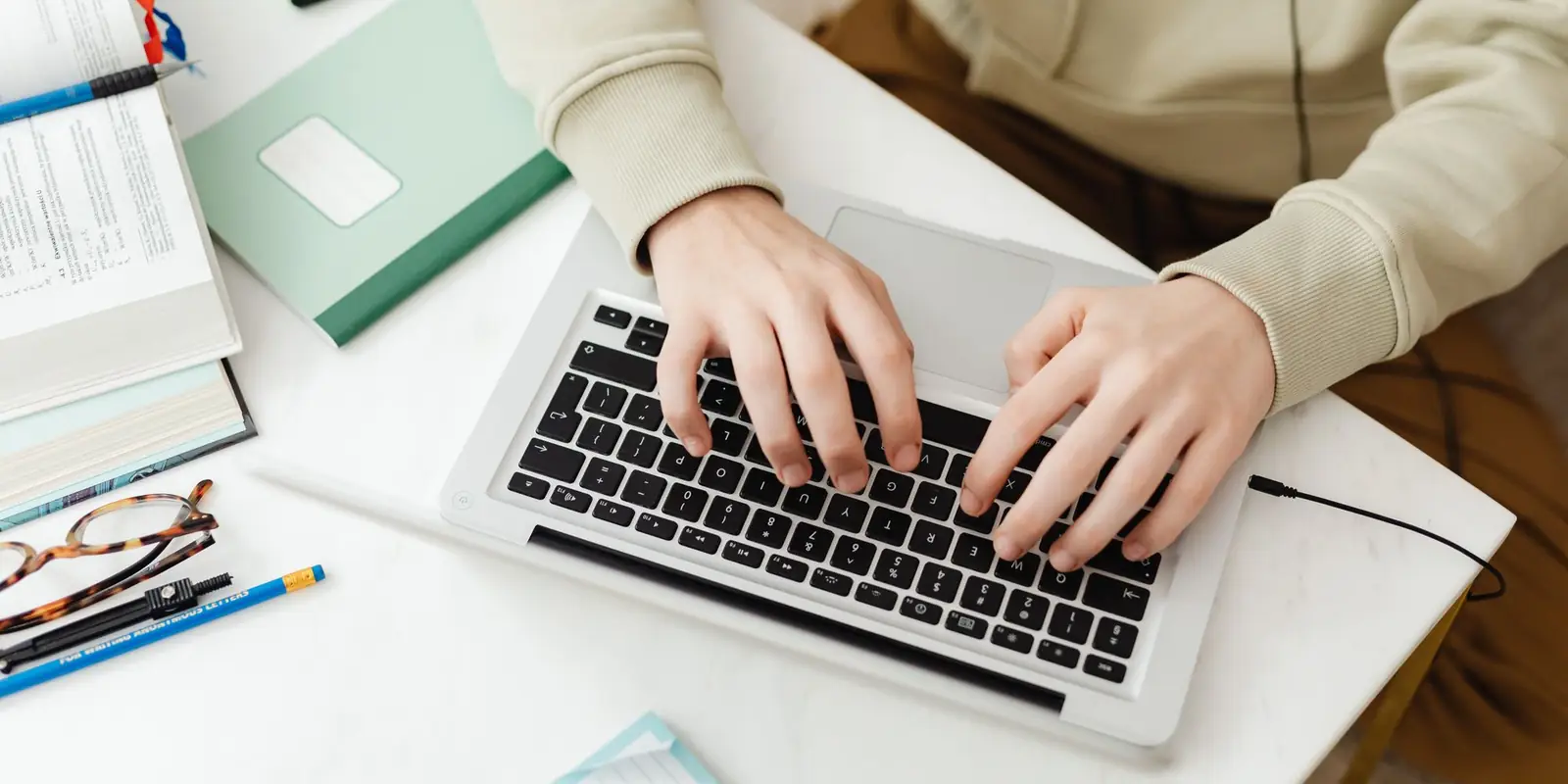Hands typing on a silver laptop on a white desk with notebooks and pens