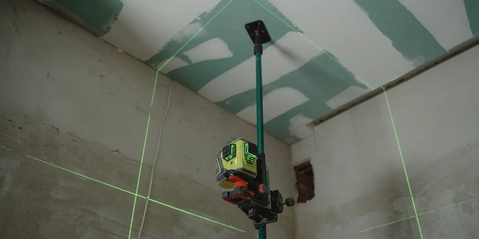 A laser level on a pole projecting green lines across the walls of an unfinished room