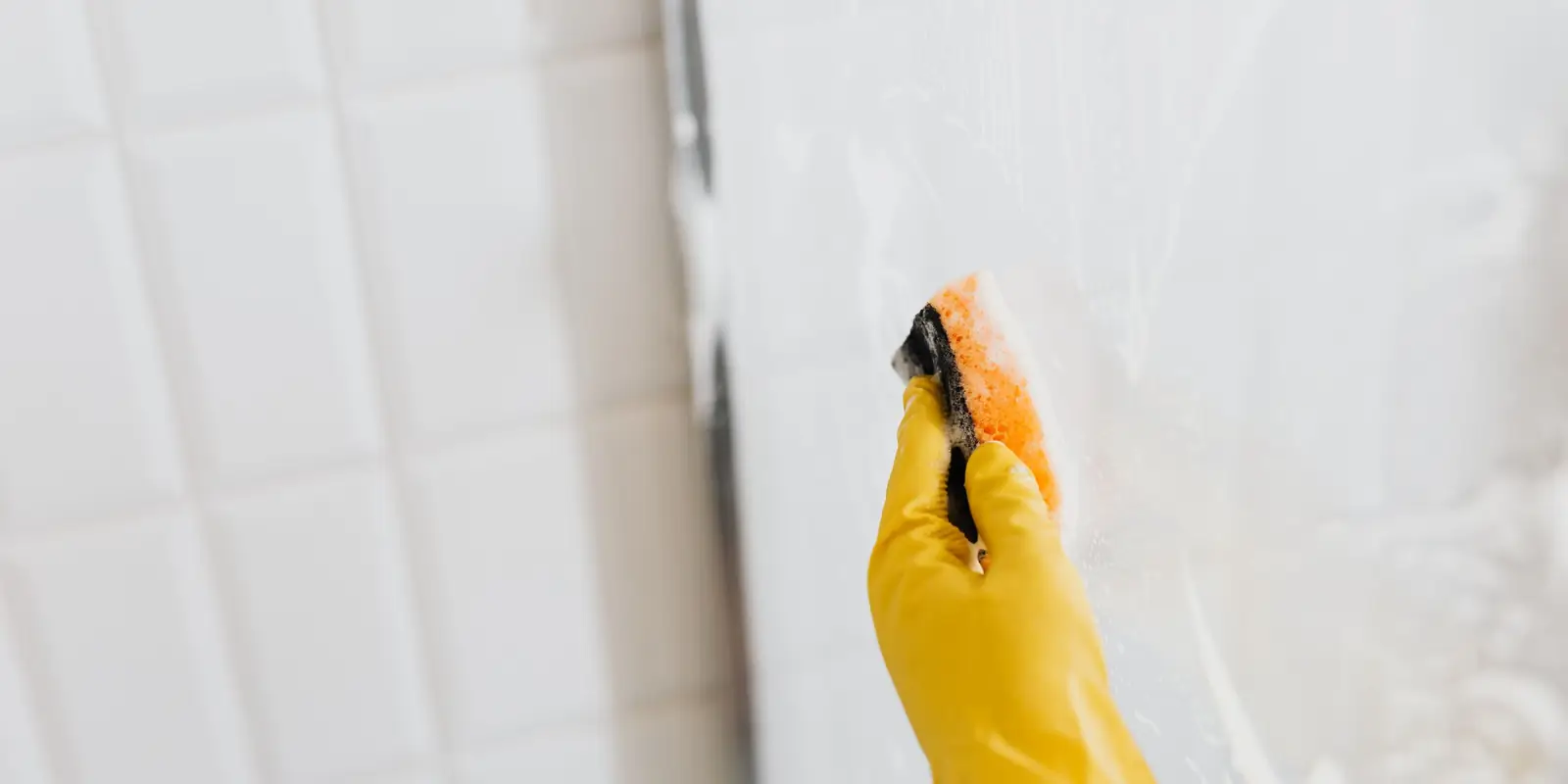 A hand in a yellow rubber glove cleaning a glass shower screen with a sponge