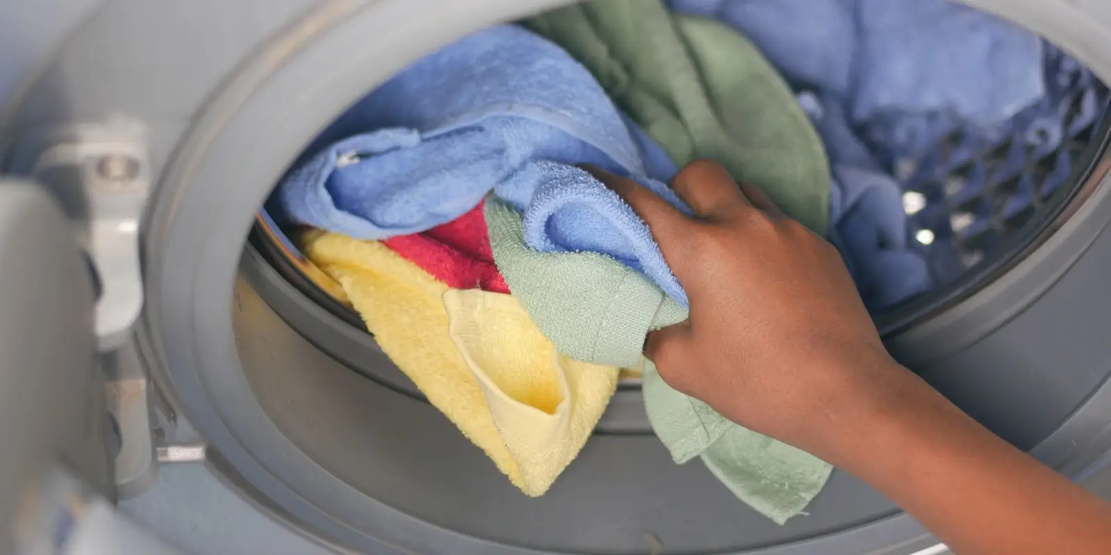 A hand loading coloured towels into a front-loading washing machine with its rubber door seal visible