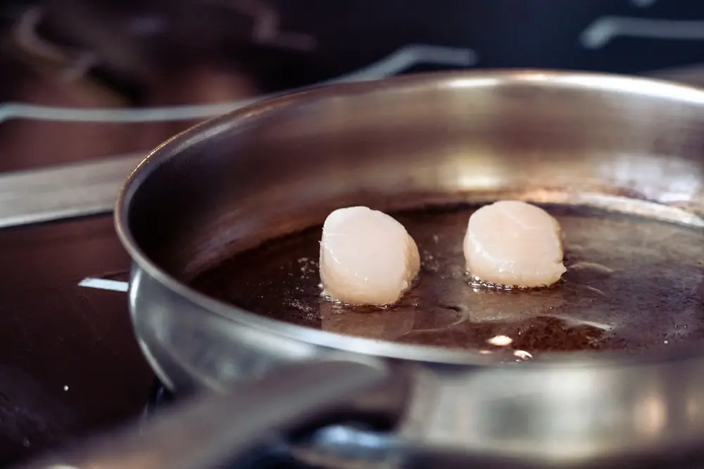 Two scallops searing in a stainless steel frying pan on a hob