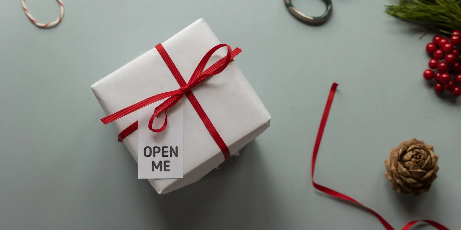 A white gift box tied with red ribbon and an "Open me" tag, with scissors, pine cones and berries