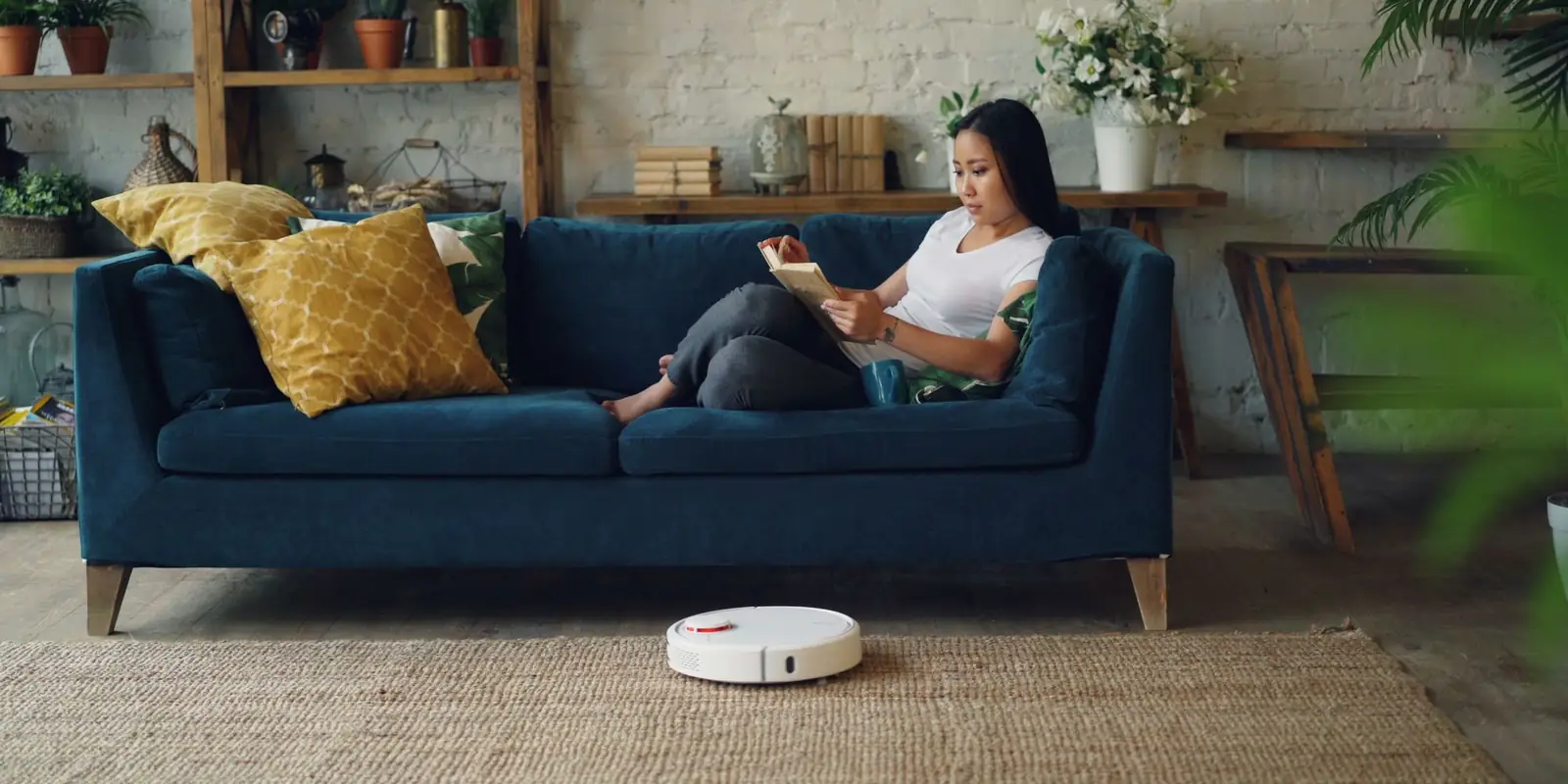 Woman reading on a sofa while a robot vacuum cleans the living room floor