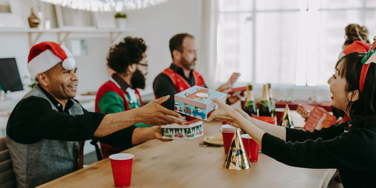 Colleagues in Santa hats and festive jumpers swapping wrapped presents across an office table