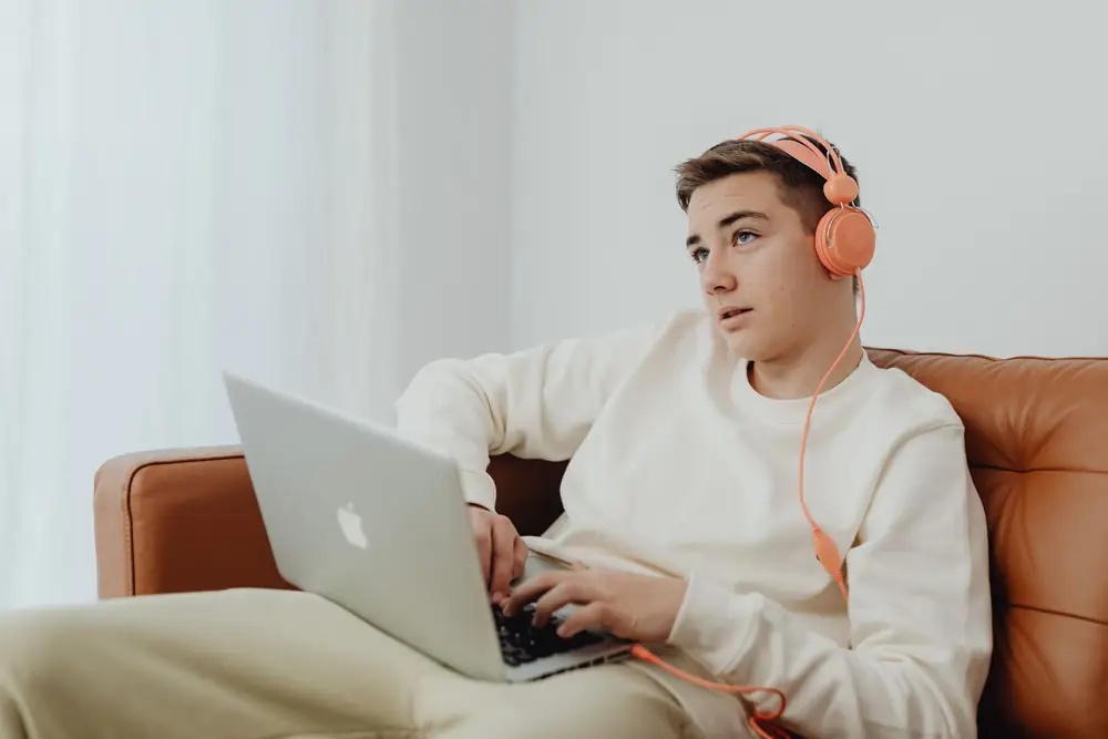 Teenage boy wearing orange headphones and using a laptop on a sofa