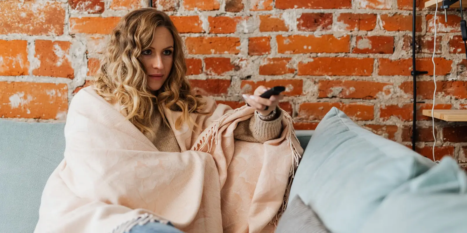 Woman wrapped in a cream blanket on a sofa holding a TV remote