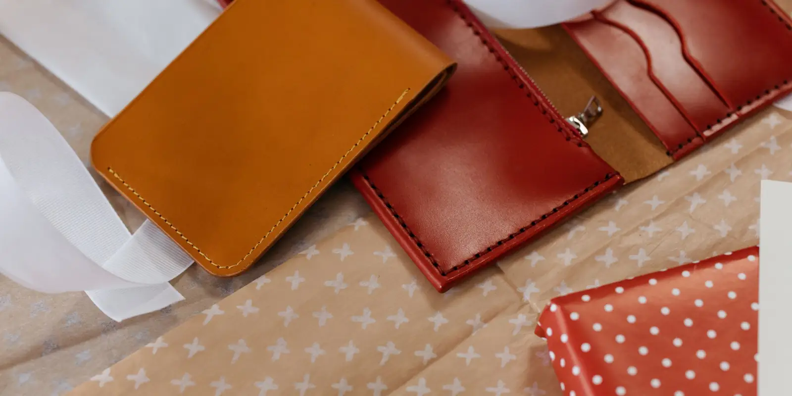 A tan leather wallet and an open red leather wallet on spotted gift wrap with white ribbon
