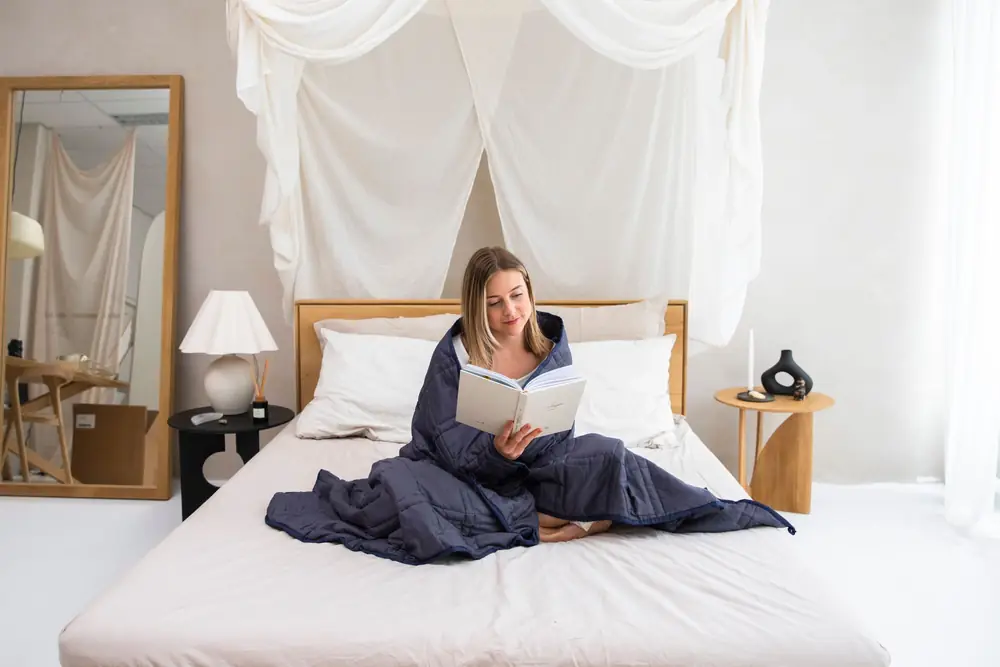 Woman sitting up in bed reading a book with a dark weighted blanket over her legs